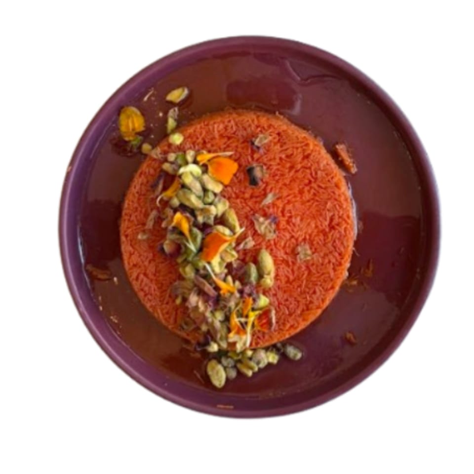 A vibrant orange Middle Eastern dessert, likely kunafa, garnished with pistachios and edible flowers on a purple plate.